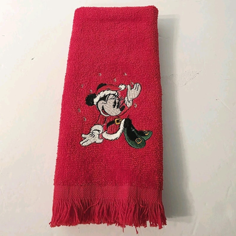 Puede incluir: Toalla de mano roja con una imagen bordada de Minnie Mouse con un traje de Pap&aacute; Noel. La toalla tiene un borde con flecos y est&aacute; hecha de un material suave y absorbente. El dise&ntilde;o de Minnie Mouse es en negro, blanco, rojo y verde.