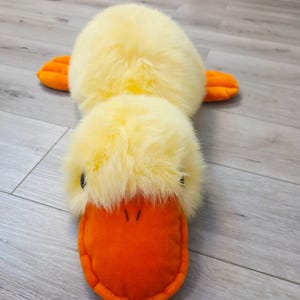 Pode incluir: Um brinquedo de pelúcia em forma de pato com corpo e cabeça de pelo amarelo brilhante e um bico laranja liso e vibrante. O brinquedo tem dois olhos pequenos, escuros e redondos, e duas patas palmadas laranja planas nas laterais do corpo.