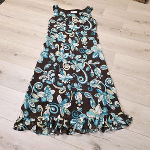 May include: A sleeveless, ankle-length dress in a dark brown color featuring a repeating floral and swirl pattern in shades of light blue, white, and pale yellow. The garment has a round neckline and a ruffled hemline at the bottom edge.