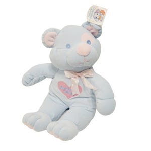 May include: A light blue teddy bear with pink detailing on the ears, nose, and a heart-shaped patch on the chest. The bear has a pink bow and the text "Baby's First" on the heart. A tag is attached to the ear.