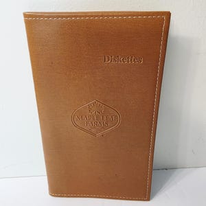 May include: A brown leatherette holder with white stitching. The text "Diskettes" is embossed at the top, and a Maple Leaf Farms logo is centered below. The holder has a slightly angled design.
