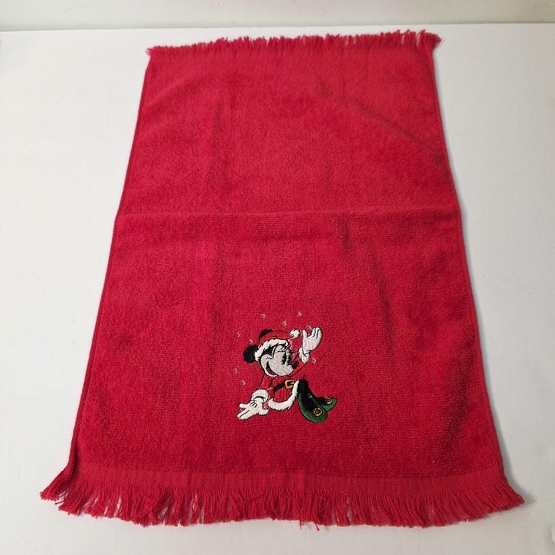 Puede incluir: Una toalla de mano roja con borde con flecos. Una imagen bordada de Mickey Mouse con un traje de Pap&aacute; Noel aparece en la toalla. La toalla est&aacute; hecha de un material suave y absorbente, ideal para el ba&ntilde;o o la cocina.