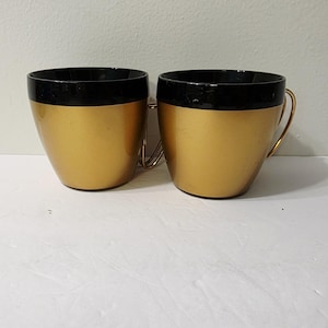 Op de afbeelding: Twee bijpassende mokken met een metallic gouden afwerking aan de onderkant en een glanzende zwarte band langs de bovenrand. Elke mok is voorzien van een dun, gebogen goudkleurig metalen handvat aan de zijkant voor een uniforme uitstraling.