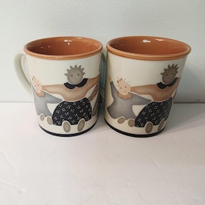 May include: Two ceramic mugs with a cream-colored exterior and a brown interior. Each mug features a whimsical illustration of three figures holding hands. The figures are dressed in gray, brown, and black clothing. The mugs have a white handle and a black base.