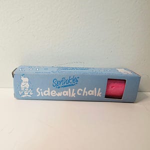 May include: A light blue rectangular cardboard box containing a bright pink stick of sidewalk chalk. The packaging features the text Sprinkles of Ponderosa Sidewalk Chalk, a cartoon character illustration, and small white rectangular graphic accents.