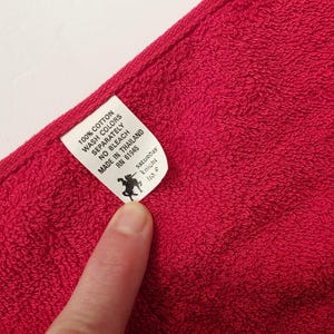 Puede incluir: Primer plano de una toalla de algod&oacute;n roja vibrante. Una etiqueta blanca muestra el texto: "100% Algod&oacute;n, Lavar los colores por separado, No usar lej&iacute;a, Hecho en Tailandia, RN 61945." La etiqueta tambi&eacute;n presenta un logotipo negro y las palabras "saturday knight ltd."