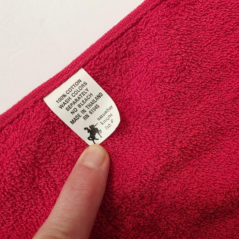 Puede incluir: Primer plano de una toalla de algod&oacute;n roja vibrante. Una etiqueta blanca muestra el texto: "100% Algod&oacute;n, Lavar los colores por separado, No usar lej&iacute;a, Hecho en Tailandia, RN 61945." La etiqueta tambi&eacute;n presenta un logotipo negro y las palabras "saturday knight ltd."