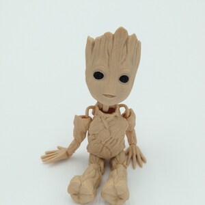 May include: A tan-colored figurine of a character with a tree-like appearance, featuring a head with leaf-like structures, black eyes, and a small smile. The figurine is seated with arms and legs extended, showcasing detailed textures.