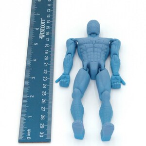 May include: A light blue action figure of a superhero, approximately 6 inches tall, with a matching ruler for scale. The figure has articulated joints and is posed upright. The ruler is marked in inches.