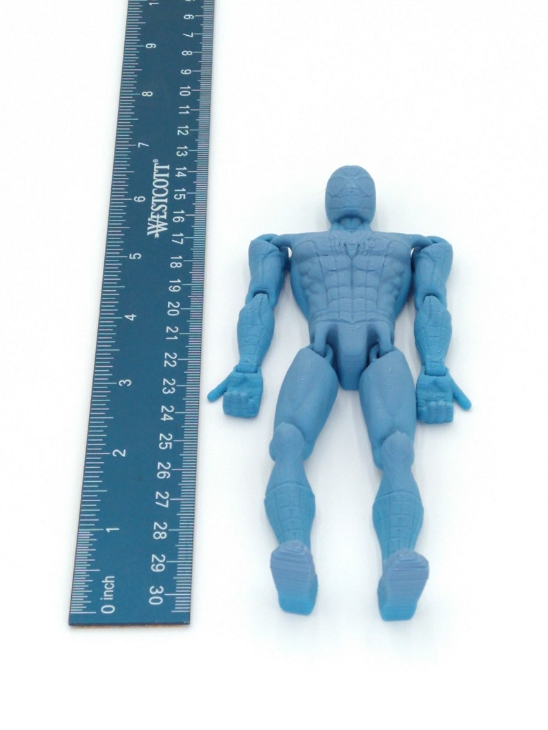 May include: A light blue action figure of a superhero, approximately 6 inches tall, with a matching ruler for scale. The figure has articulated joints and is posed upright. The ruler is marked in inches.