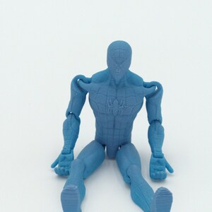 May include: A light blue action figure of a superhero, seated with legs extended. The figure has a textured surface and visible joints at the shoulders, elbows, hips, and knees. A spider emblem is visible on the chest.