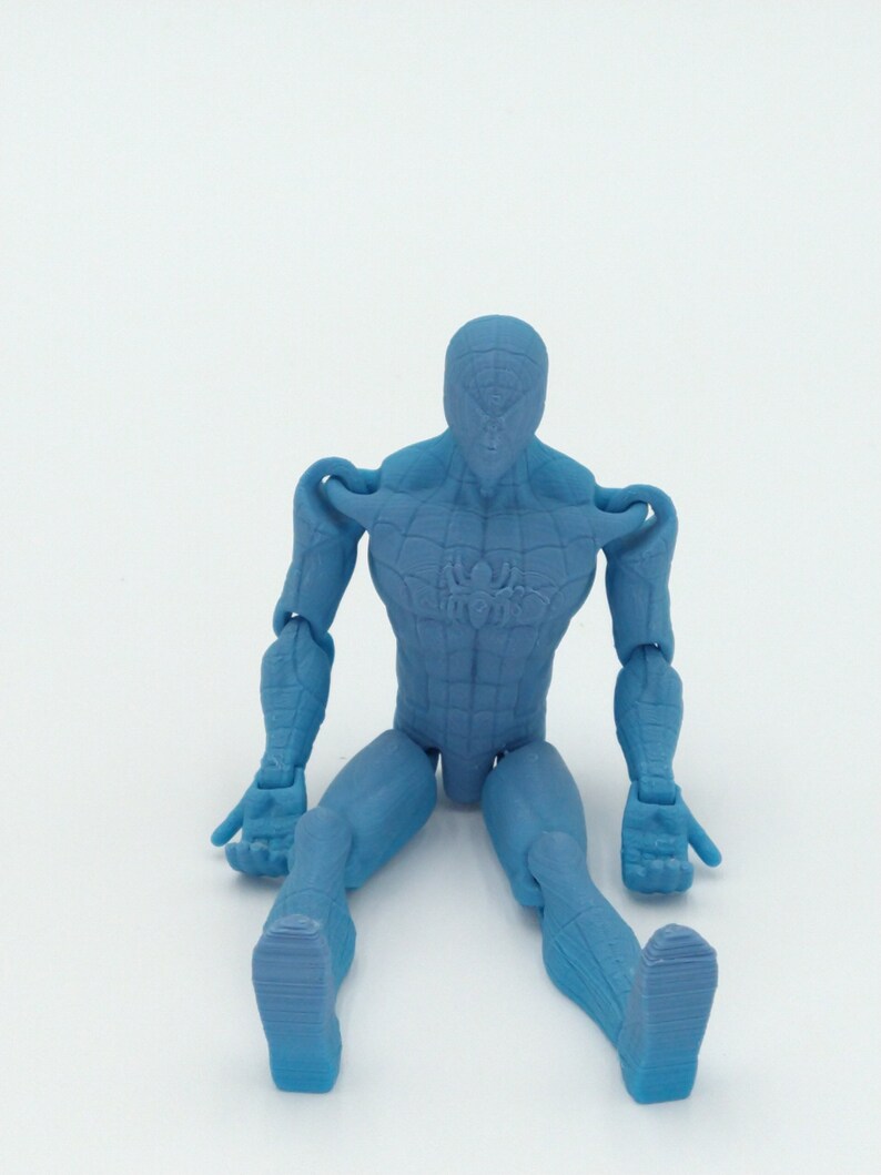 May include: A light blue action figure of a superhero, seated with legs extended. The figure has a textured surface and visible joints at the shoulders, elbows, hips, and knees. A spider emblem is visible on the chest.