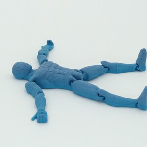 May include: A blue articulated action figure in a prone position, lying on its back with arms and legs spread. The figure has a detailed design, with visible joints and sculpted features. The figure is made of a solid, opaque material.