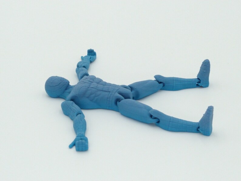 May include: A blue articulated action figure in a prone position, lying on its back with arms and legs spread. The figure has a detailed design, with visible joints and sculpted features. The figure is made of a solid, opaque material.