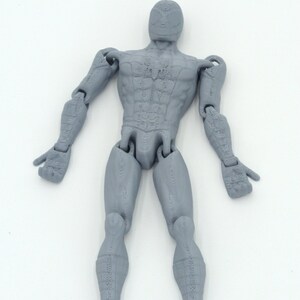 May include: A gray articulated action figure of a human form, with detailed musculature and jointed limbs. The figure is posed with arms outstretched and legs slightly apart, showcasing its range of motion. The figure is made of a solid gray material.