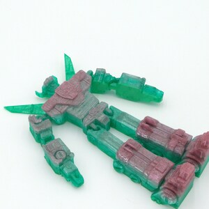 May include: A disassembled, translucent robot toy with a mix of green and purple hues. The toy is lying on a white surface, showcasing its articulated limbs and body parts. The plastic robot appears to be a model kit or a collectible item.