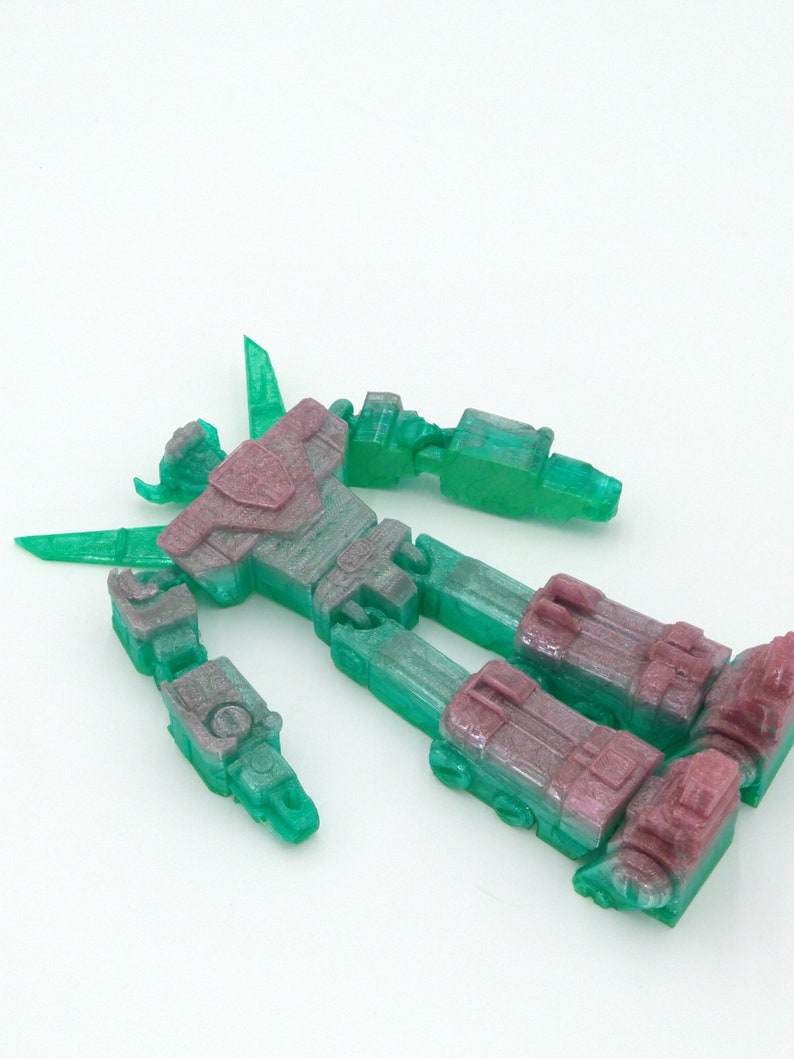 May include: A disassembled, translucent robot toy with a mix of green and purple hues. The toy is lying on a white surface, showcasing its articulated limbs and body parts. The plastic robot appears to be a model kit or a collectible item.