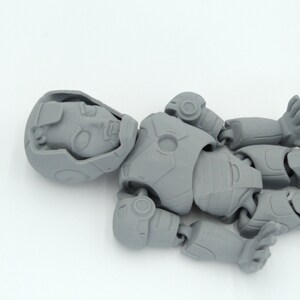 May include: A gray, articulated action figure, lying on a white surface. The figure is in a robotic or futuristic style, with visible joints and detailed armor-like plating. The head is detailed with facial features.