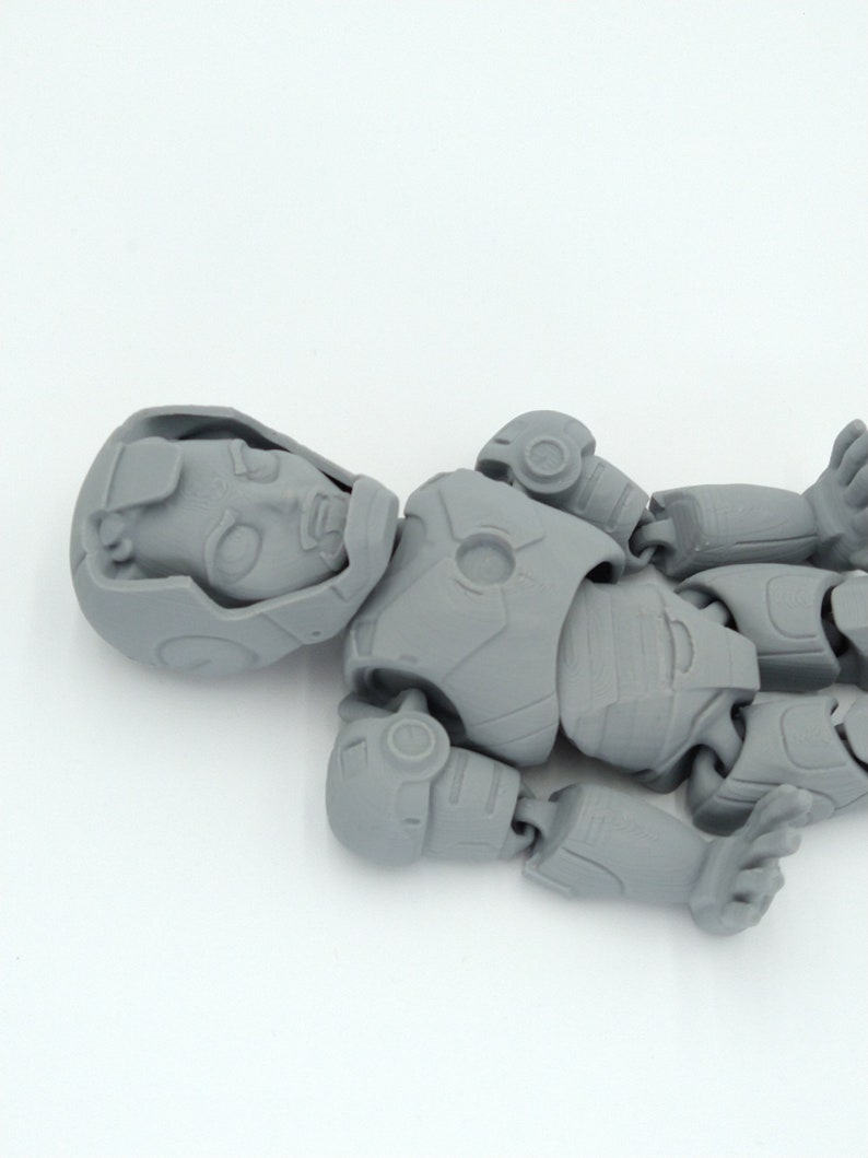 May include: A gray, articulated action figure, lying on a white surface. The figure is in a robotic or futuristic style, with visible joints and detailed armor-like plating. The head is detailed with facial features.
