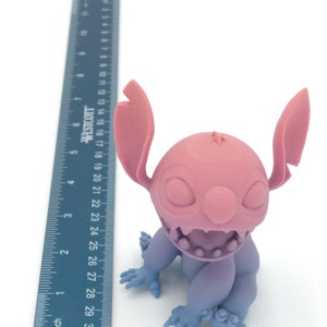 Puede incluir: Una figura impresa en 3D de un personaje de dibujos animados, con cabeza y orejas rosas, cuerpo morado claro y manos y pies azules. La figura est&aacute; junto a una regla que mide hasta 76 cm.