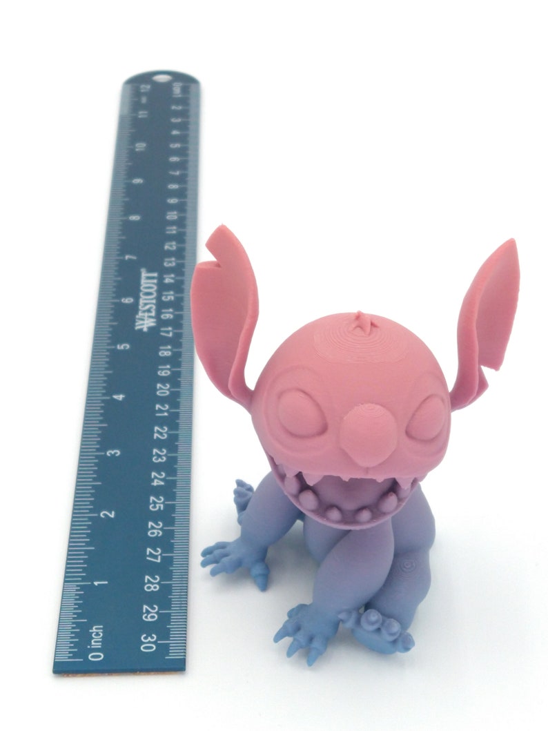 Puede incluir: Una figura impresa en 3D de un personaje de dibujos animados, con cabeza y orejas rosas, cuerpo morado claro y manos y pies azules. La figura est&aacute; junto a una regla que mide hasta 76 cm.