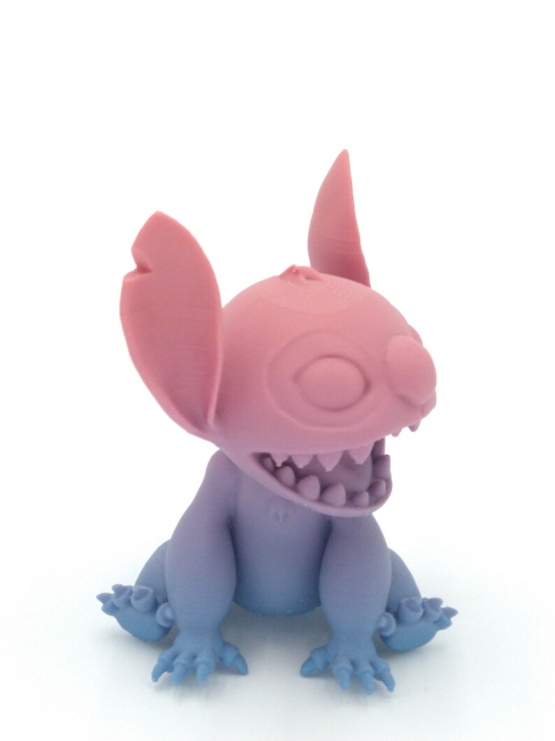 Puede incluir: Figura de dos tonos de un personaje alien&iacute;gena de dibujos animados. La cabeza y las orejas son rosas, mientras que el cuerpo y las extremidades hacen la transici&oacute;n de azul a morado. La figura tiene una boca muy abierta con dientes visibles y ojos grandes y expresivos.