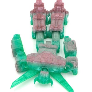 May include: A disassembled, translucent toy robot in shades of green, pink, and gray. The robot is composed of multiple geometric parts, including two upright sections, a central body, and wing-like extensions. The pieces are arranged on a white background.