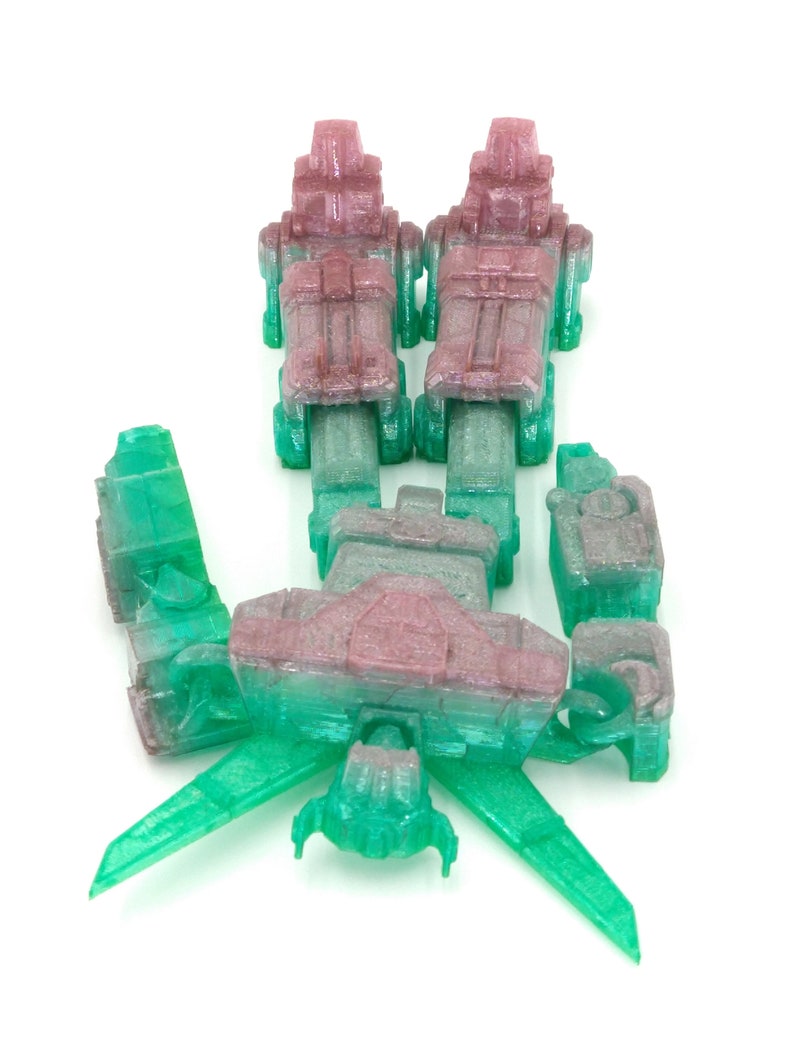 May include: A disassembled, translucent toy robot in shades of green, pink, and gray. The robot is composed of multiple geometric parts, including two upright sections, a central body, and wing-like extensions. The pieces are arranged on a white background.