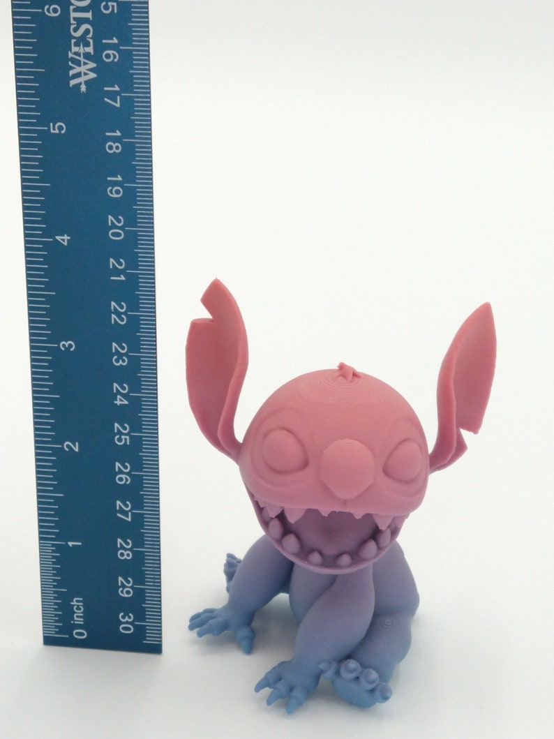 Puede incluir: Figura impresa en 3D de un personaje de dibujos animados, con cabeza y orejas rosas, cara y cuerpo morados, y manos y pies azules. La figura mide aproximadamente 7,6 cm de altura. El personaje tiene una amplia sonrisa dentada.