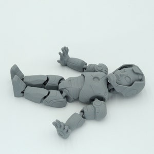 May include: A disassembled gray action figure of a humanoid robot, lying on a white surface. The figure is segmented at the joints, with visible details on the head, torso, arms, and legs. The figure is in a relaxed pose.
