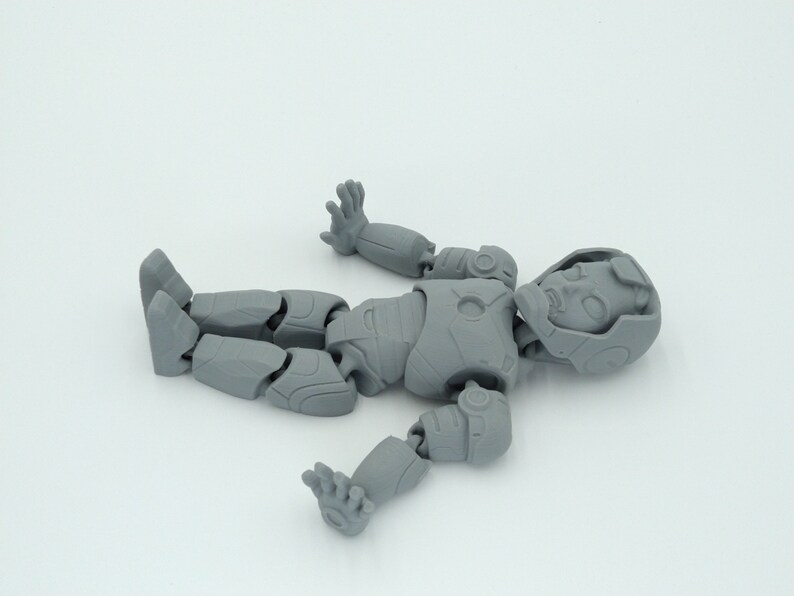 May include: A disassembled gray action figure of a humanoid robot, lying on a white surface. The figure is segmented at the joints, with visible details on the head, torso, arms, and legs. The figure is in a relaxed pose.