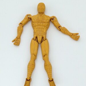 May include: A tan action figure of a superhero with articulated joints. The figure is posed with arms outstretched and legs slightly apart. The figure has detailed musculature and a textured surface.