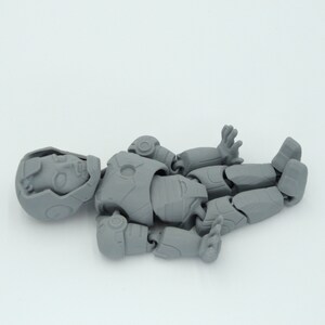 May include: A disassembled gray action figure, lying on a white surface. The figure is in multiple pieces, including the head, torso, arms, and legs. The figure has a detailed design, with visible joints and armor-like features.