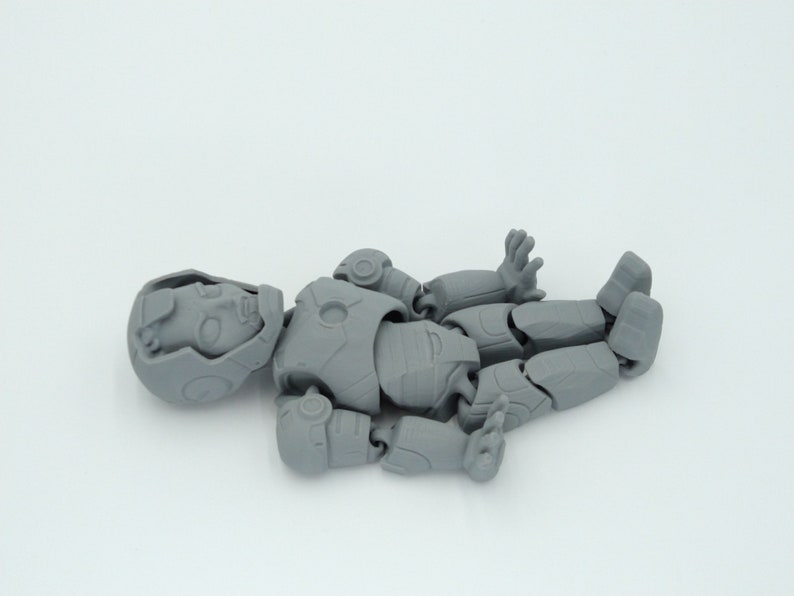 May include: A disassembled gray action figure, lying on a white surface. The figure is in multiple pieces, including the head, torso, arms, and legs. The figure has a detailed design, with visible joints and armor-like features.
