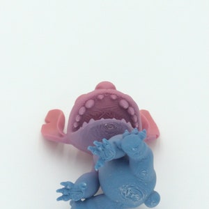 Puede incluir: Una figura azul y morada de un personaje de dibujos animados, tumbada boca arriba. La figura tiene orejas grandes, una boca ancha con dientes visibles y cuatro brazos y piernas. Los colores son una mezcla de azul, morado y rosa.