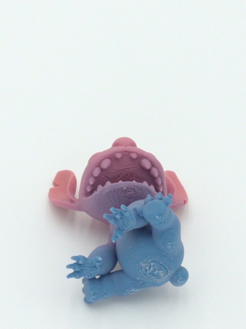 Puede incluir: Una figura azul y morada de un personaje de dibujos animados, tumbada boca arriba. La figura tiene orejas grandes, una boca ancha con dientes visibles y cuatro brazos y piernas. Los colores son una mezcla de azul, morado y rosa.