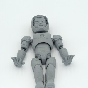 May include: A gray, articulated action figure of a superhero, featuring a detailed helmet, chest plate, and jointed limbs. The figure is posed with arms outstretched and hands open, showcasing its design. The toy is made of a solid, matte material.