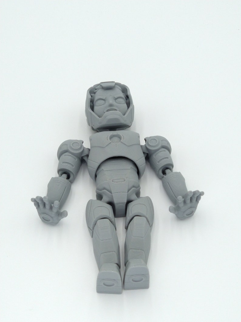 May include: A gray, articulated action figure of a superhero, featuring a detailed helmet, chest plate, and jointed limbs. The figure is posed with arms outstretched and hands open, showcasing its design. The toy is made of a solid, matte material.