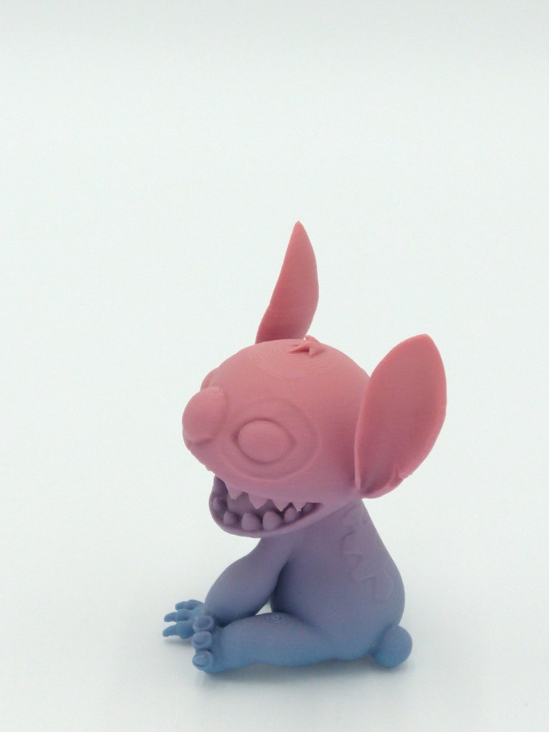 Puede incluir: Una figura rosa y azul de un personaje de dibujos animados con orejas grandes, una amplia sonrisa y cuatro dedos. La figura est&aacute; sentada con los brazos y las piernas doblados. Los colores hacen la transici&oacute;n de rosa a azul.