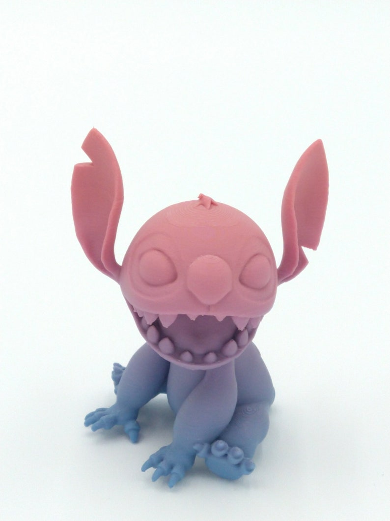 Puede incluir: Figura impresa en 3D de un personaje de dibujos animados, con cabeza y orejas rosas, boca muy abierta y cuerpo y extremidades azul-violeta. La figura est&aacute; sentada, con las manos y los pies visibles.