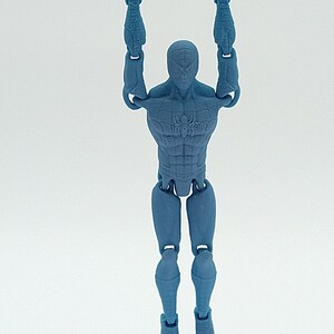 May include: A blue action figure of a superhero hanging from a white bar. The figure has a detailed design, with visible muscles and a spider emblem on the chest. The figure is suspended in mid-air against a white background.