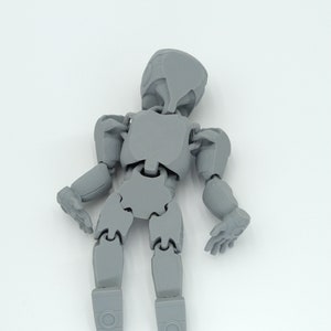 May include: A gray articulated figure with a rounded head, jointed limbs, and hands. The figure is designed to be posed and is made of a solid, matte material. The figure is standing against a white background.