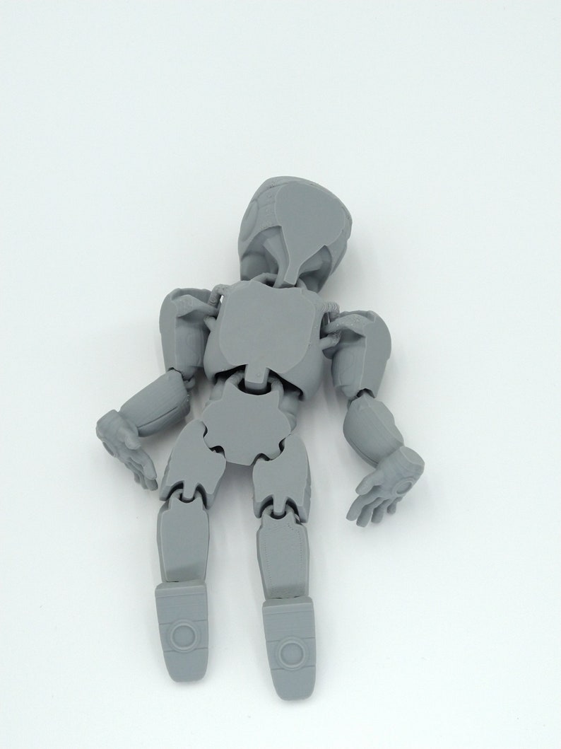 May include: A gray articulated figure with a rounded head, jointed limbs, and hands. The figure is designed to be posed and is made of a solid, matte material. The figure is standing against a white background.