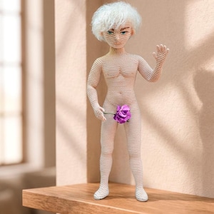 May include: A crocheted doll with a light skin tone, white hair, and blue eyes. The doll is nude, with a purple flower covering the genital area. It stands on a wooden shelf, with one hand raised in a wave. The background is a light beige.