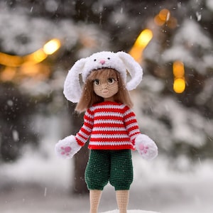 Puede incluir: Una muñeca de crochet que lleva un gorro de conejo blanco con orejas caídas, un suéter a rayas rojas y blancas, pantalones verdes y guantes blancos con huellas de patas rosas. La muñeca está de pie sobre una superficie nevada.