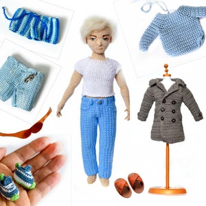 Puede incluir: Una muñeca con camiseta blanca y vaqueros azules. Otros artículos de ganchillo incluyen un suéter azul, pantalones cortos, un abrigo y zapatos. Accesorios: sandalias marrones y un objeto de madera.