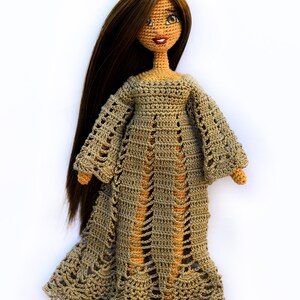 K&ouml;nnte beinhalten: Handgefertigte H&auml;kelpuppe mit langen braunen Haaren und einem beigefarbenen Kleid. Das Kleid der Puppe hat ein durchbrochenes Design mit langen &Auml;rmeln und einem ausgestellten Rock. Die Puppe hat ein l&auml;chelndes Gesicht mit braunen Augen und roten Lippen.