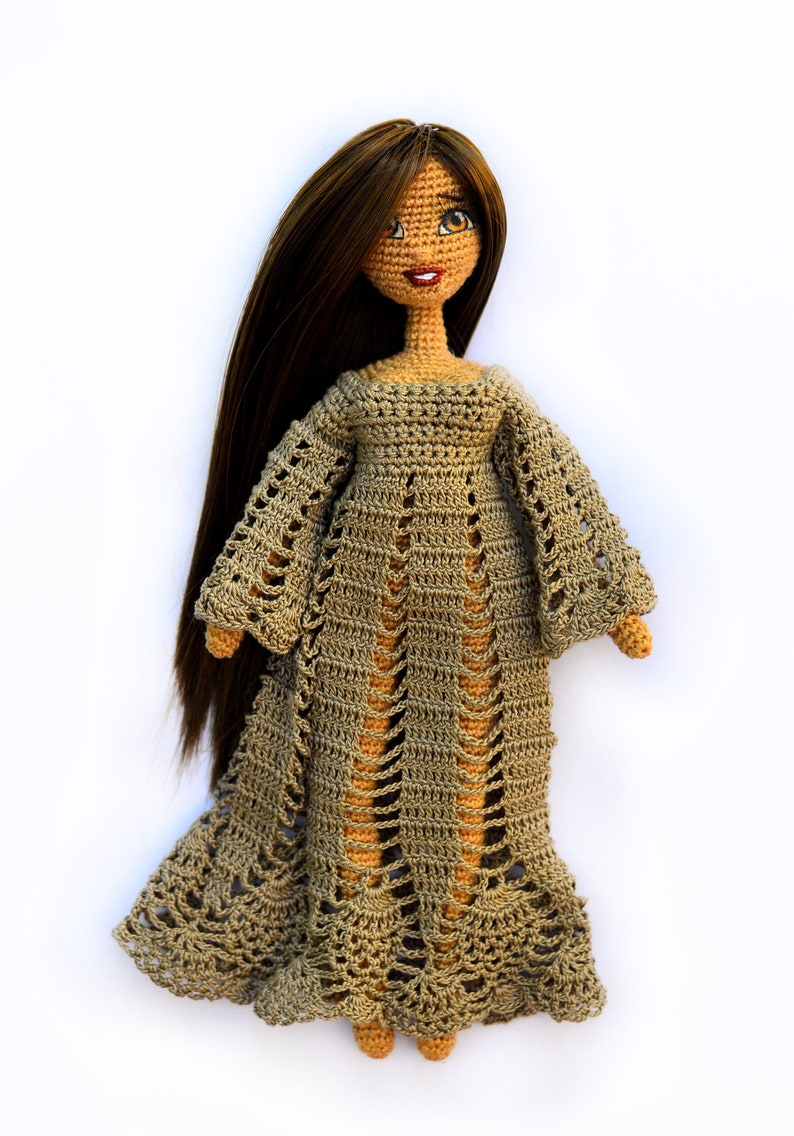 K&ouml;nnte beinhalten: Handgefertigte H&auml;kelpuppe mit langen braunen Haaren und einem beigefarbenen Kleid. Das Kleid der Puppe hat ein durchbrochenes Design mit langen &Auml;rmeln und einem ausgestellten Rock. Die Puppe hat ein l&auml;chelndes Gesicht mit braunen Augen und roten Lippen.