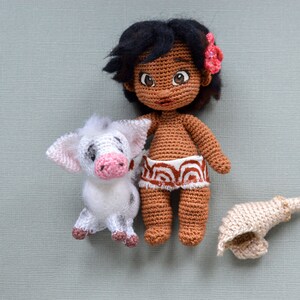 K&ouml;nnte beinhalten: Eine geh&auml;kelte Puppe mit brauner Haut, schwarzen Haaren und einer rosa Blume im Haar. Sie tr&auml;gt einen braunen und wei&szlig;en geh&auml;kelten Rock. Sie h&auml;lt ein wei&szlig;es geh&auml;keltes Schwein mit rosa Ohren und Nase. Auf dem Boden liegt eine braune geh&auml;kelte Muschel.