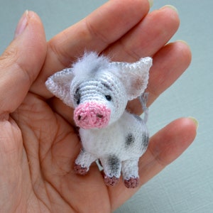 May include: A small crocheted pig toy with white and gray fur, pink ears, and a pink snout. The pig is sitting in a hand.